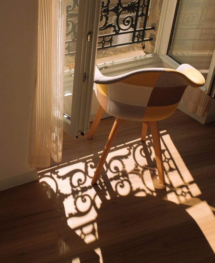 A mid-century shell chair by an open window, with low sun casting a wrought-iron pattern across a wood floor.